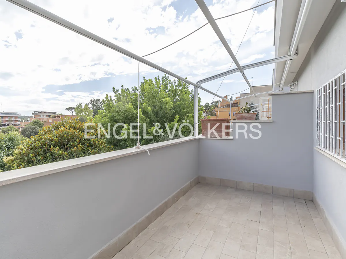 A spacious, tiled balcony with light gray walls, a metal overhead frame, and a view of lush green trees and buildings.