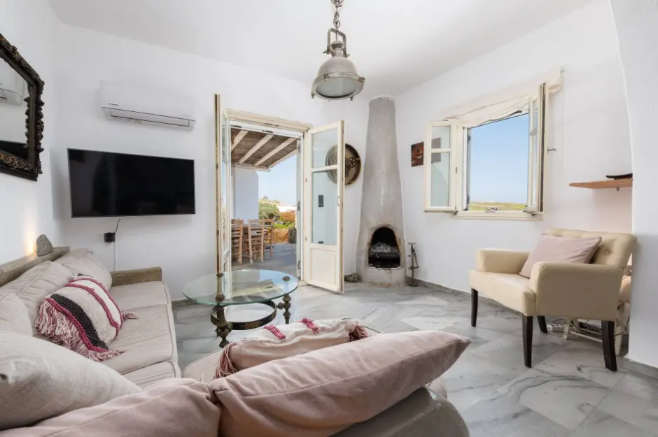 Bright living room with white walls, marble floor, beige sofa and chair, TV, and open doors to a patio with outdoor seating.