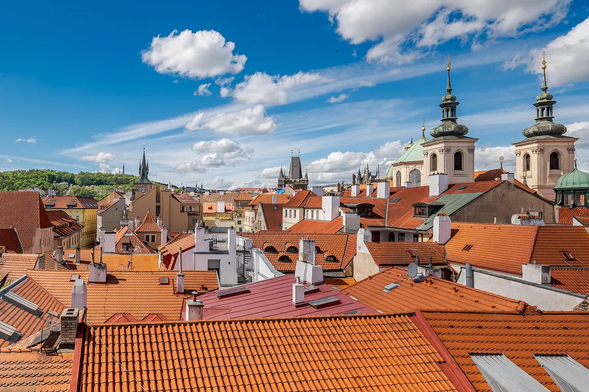 View of Prague rooftops with orange tiles under a blue sky with white clouds. Church towers rise above the buildings.