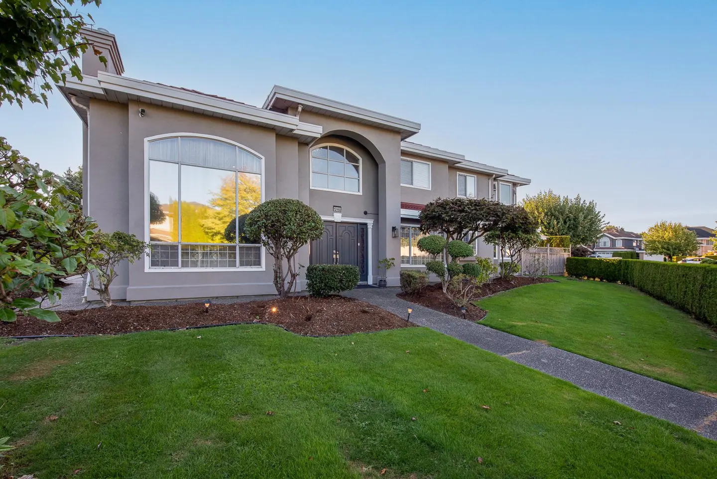 Two-story grey suburban home with a large arched window, manicured lawn, and a concrete walkway leading to the front door.