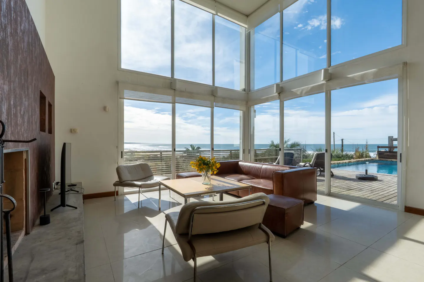 Bright living room with floor-to-ceiling windows overlooking the ocean. Brown leather sofa, chairs, and a coffee table with yellow flowers.