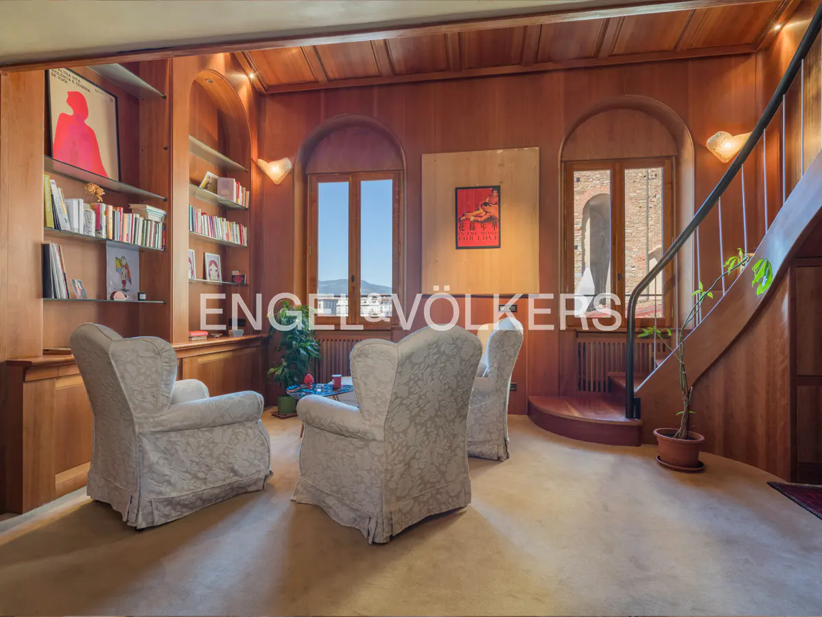 Living room with wood paneling, arched windows, and built-in bookshelves. Three floral armchairs sit on a tan carpet. A curved staircase is on the right.