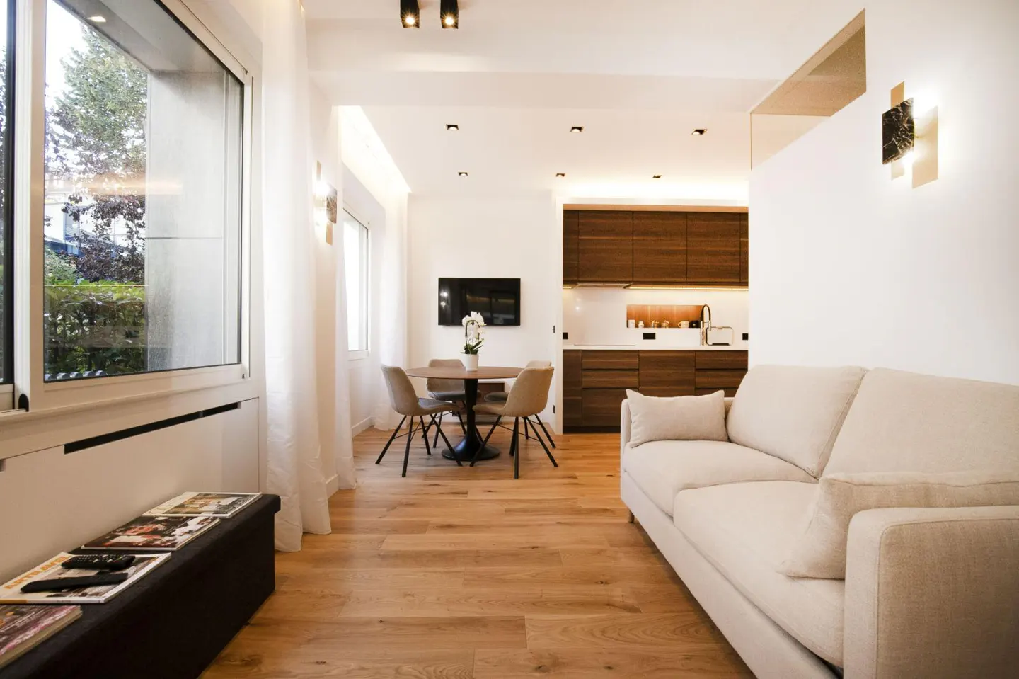 Bright, modern apartment interior with light wood floors, white walls, and a beige sofa. A round dining table sits near a window, with a kitchen in the background.