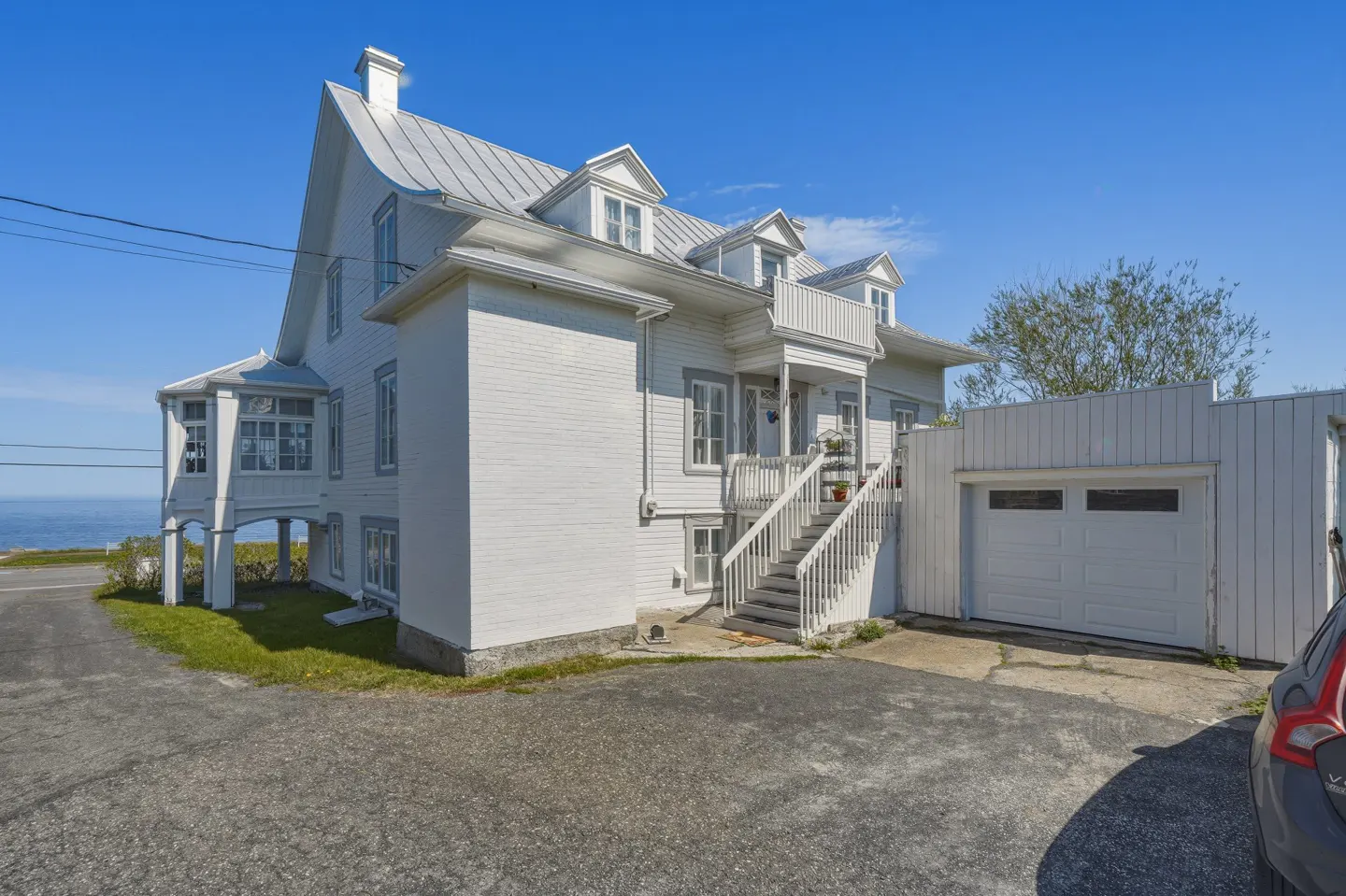 White two-story house with a metal roof, exterior staircase, and attached garage under a clear blue sky by the ocean.