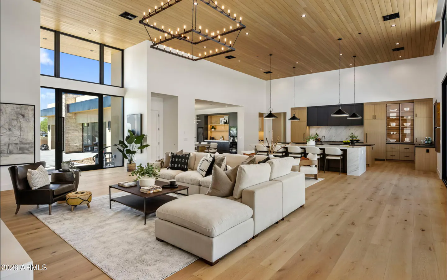 Open-concept living room with a large sectional sofa, wood floors, and a modern kitchen in the background. A large window lets in natural light.