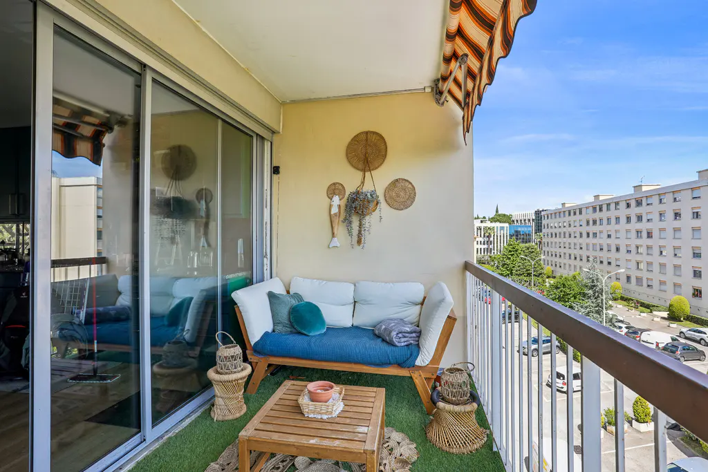 Apartment balcony with a blue couch, wooden table, and woven decor. City buildings are visible in the background.