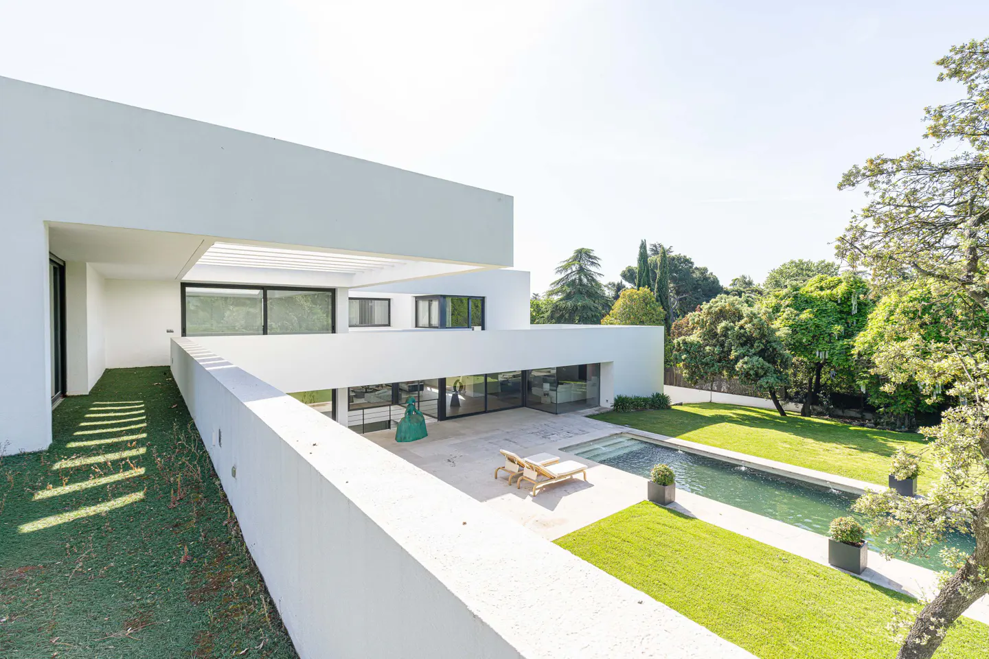 Modern white house with a pool, lawn chairs, and green lawn. Trees are in the background.