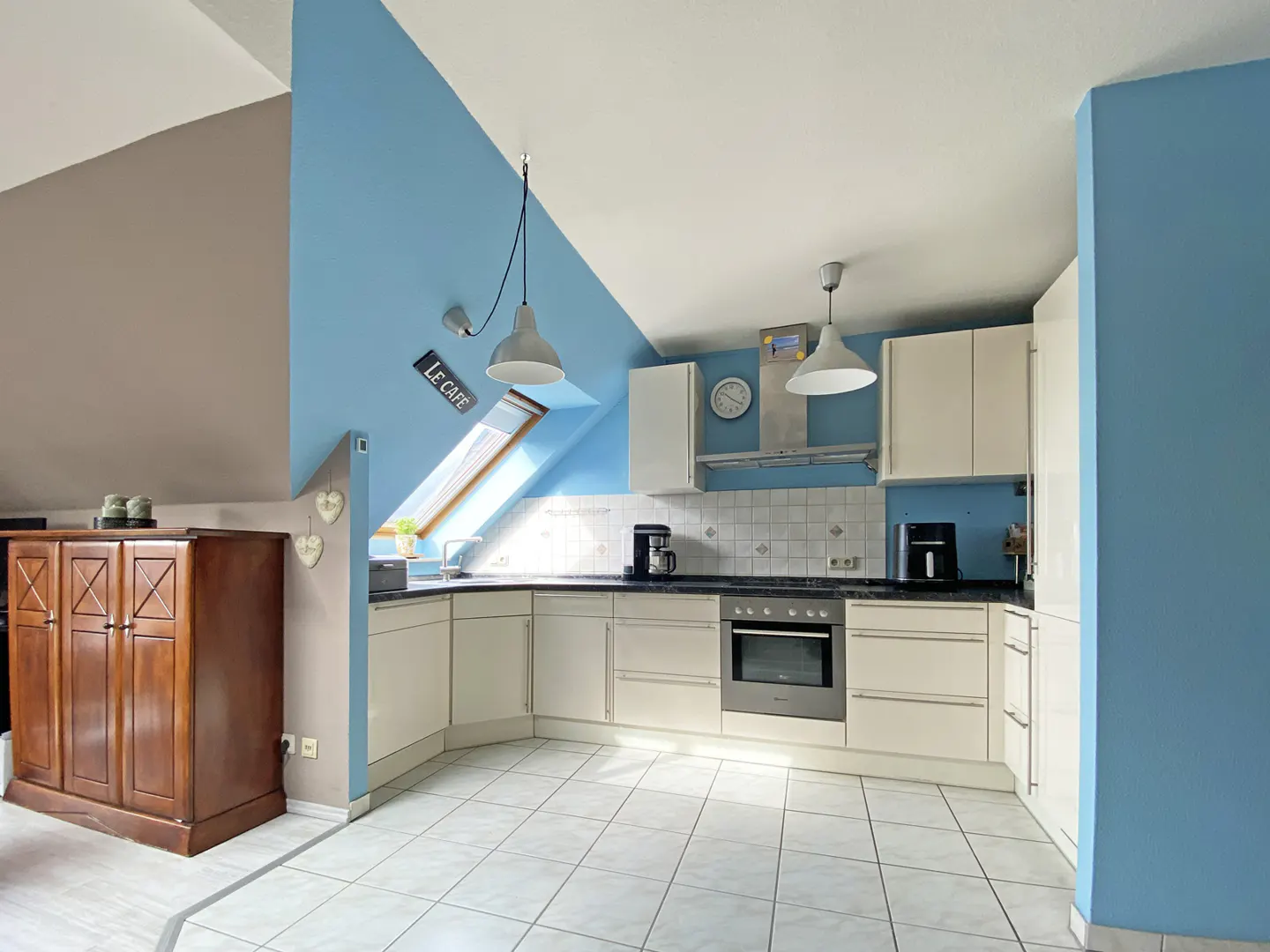 Bright kitchen with white cabinets, black countertops, and white tile floor. Blue accent wall and skylight. Brown wooden cabinet on the left.