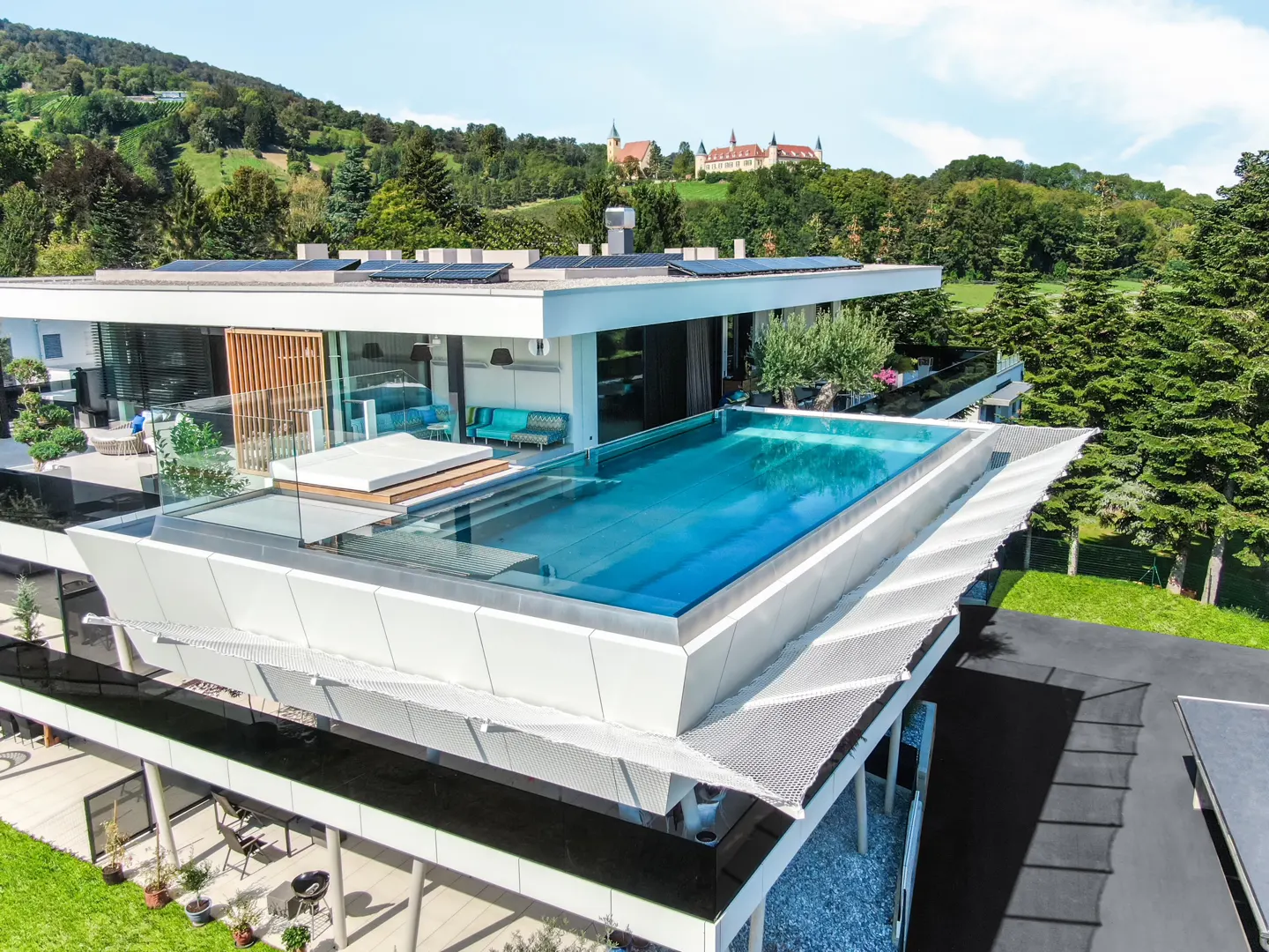 Modern white villa with a rooftop infinity pool, lounge area, and scenic views of a distant castle and green hills.