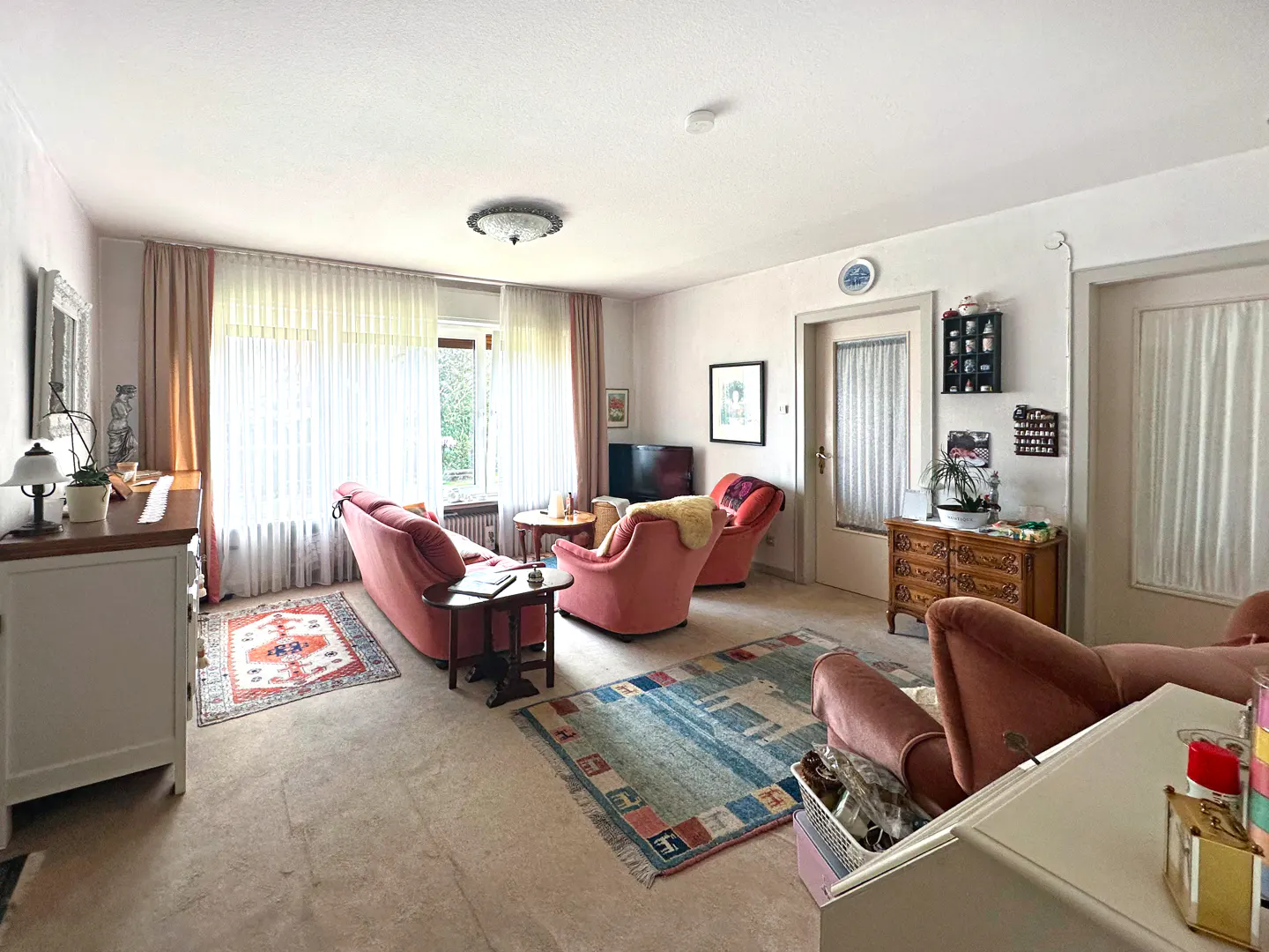 Living room with pink sofa and armchairs, patterned rugs, and white walls. Natural light filters through sheer curtains.