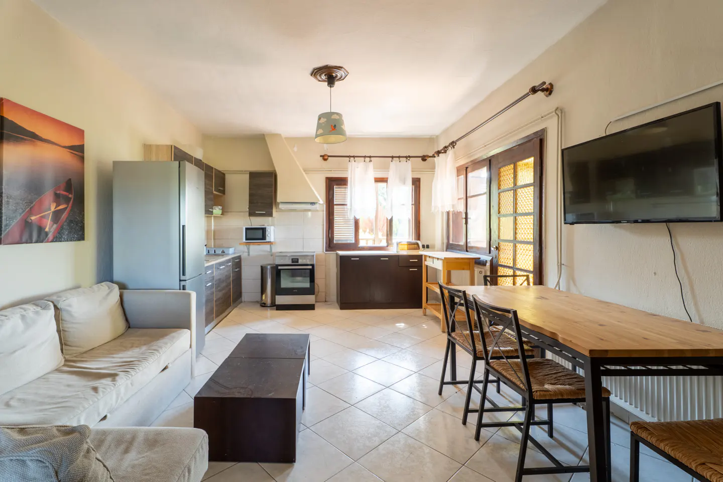 Open-concept living space with a kitchen, dining table, sofa, and TV. The kitchen has stainless steel appliances and dark wood cabinets. The dining table is wood with metal legs.