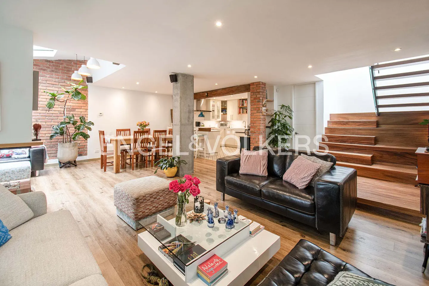 Open-concept living space with wood floors, black leather sofa, dining table, and wooden stairs. Roses on a white coffee table.