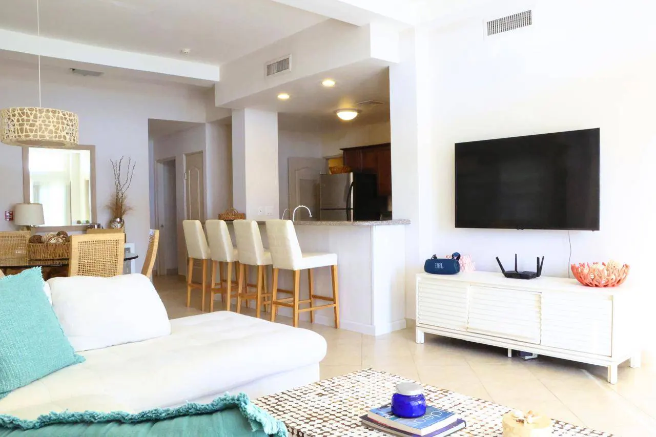 Bright, open-concept living space with white sofa, turquoise accents, and kitchen bar with four stools. A large TV hangs above a white cabinet.