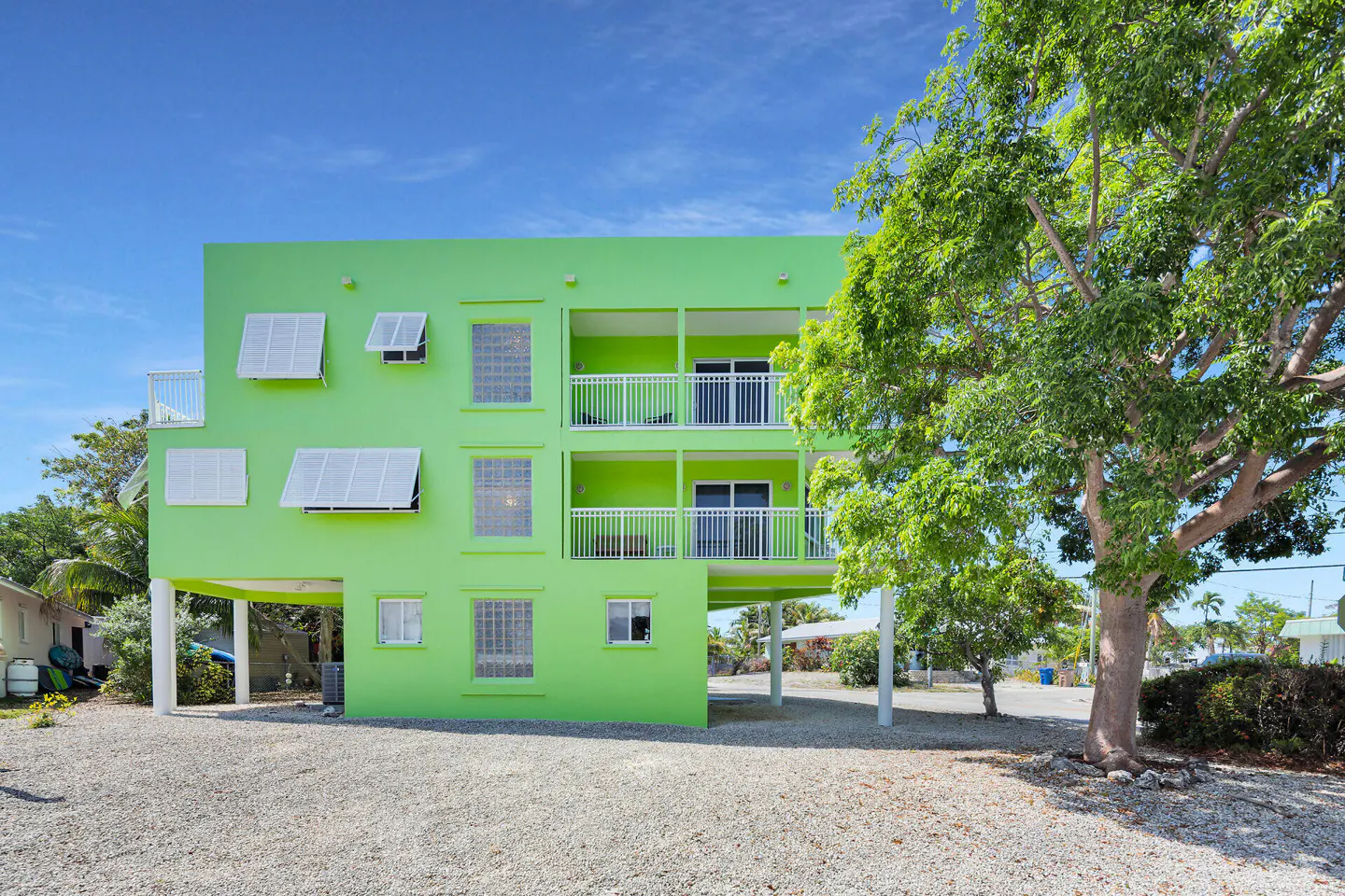 A three-story, bright green building on stilts with white trim and balconies, under a blue sky. A large tree is on the right.