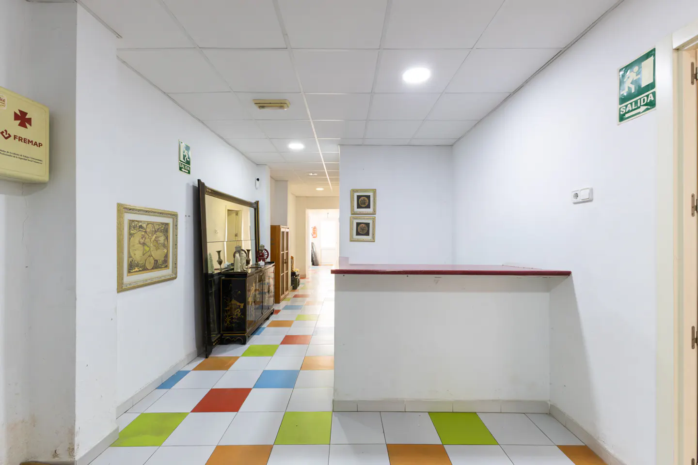 Hallway with white walls, colorful tiled floor, and a reception desk. A mirror and artwork adorn the walls. An exit sign is visible.