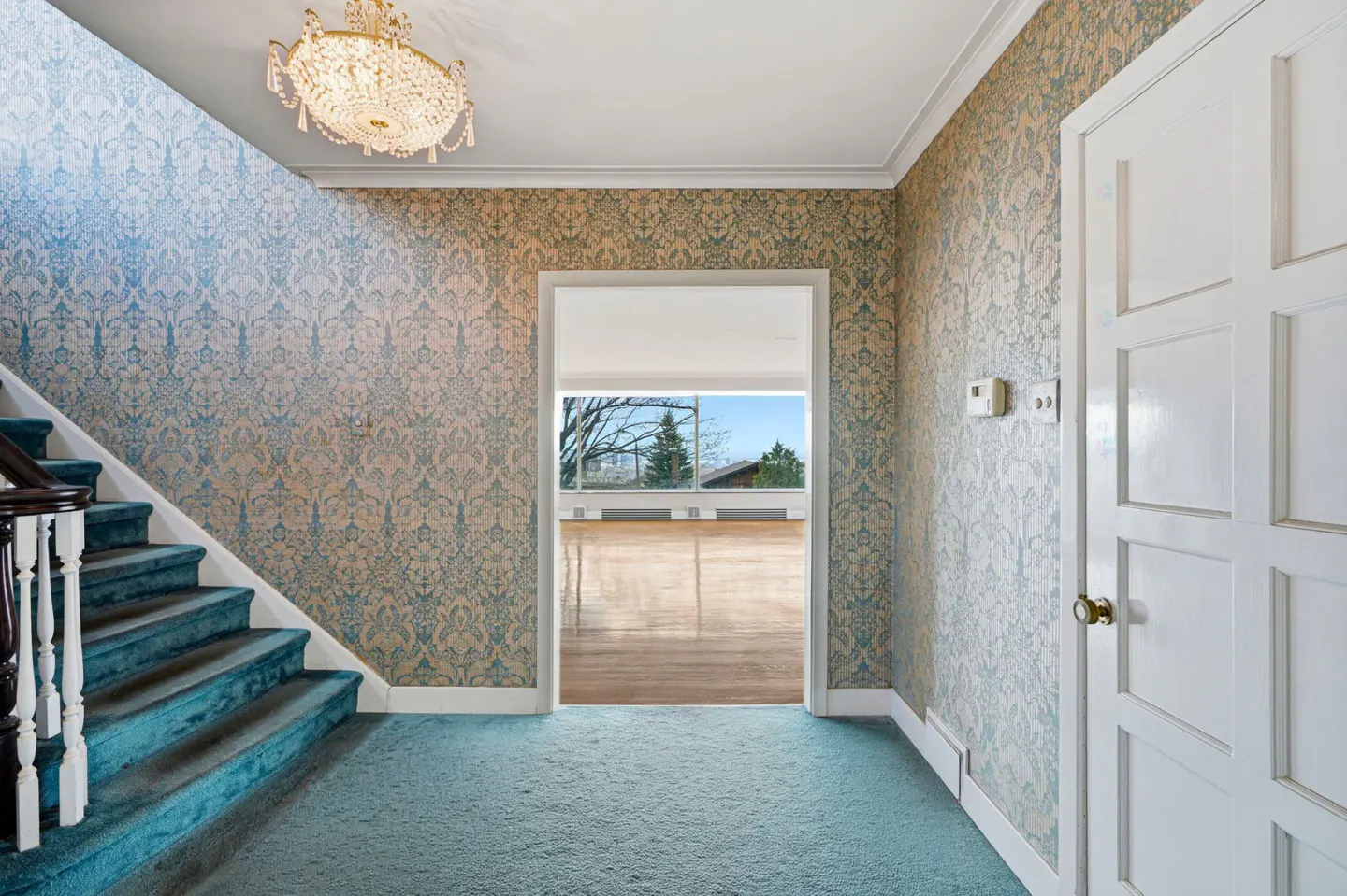 Entryway with blue carpet, patterned wallpaper, and a crystal chandelier. Stairs lead up on the left, and a doorway leads to a bright room.