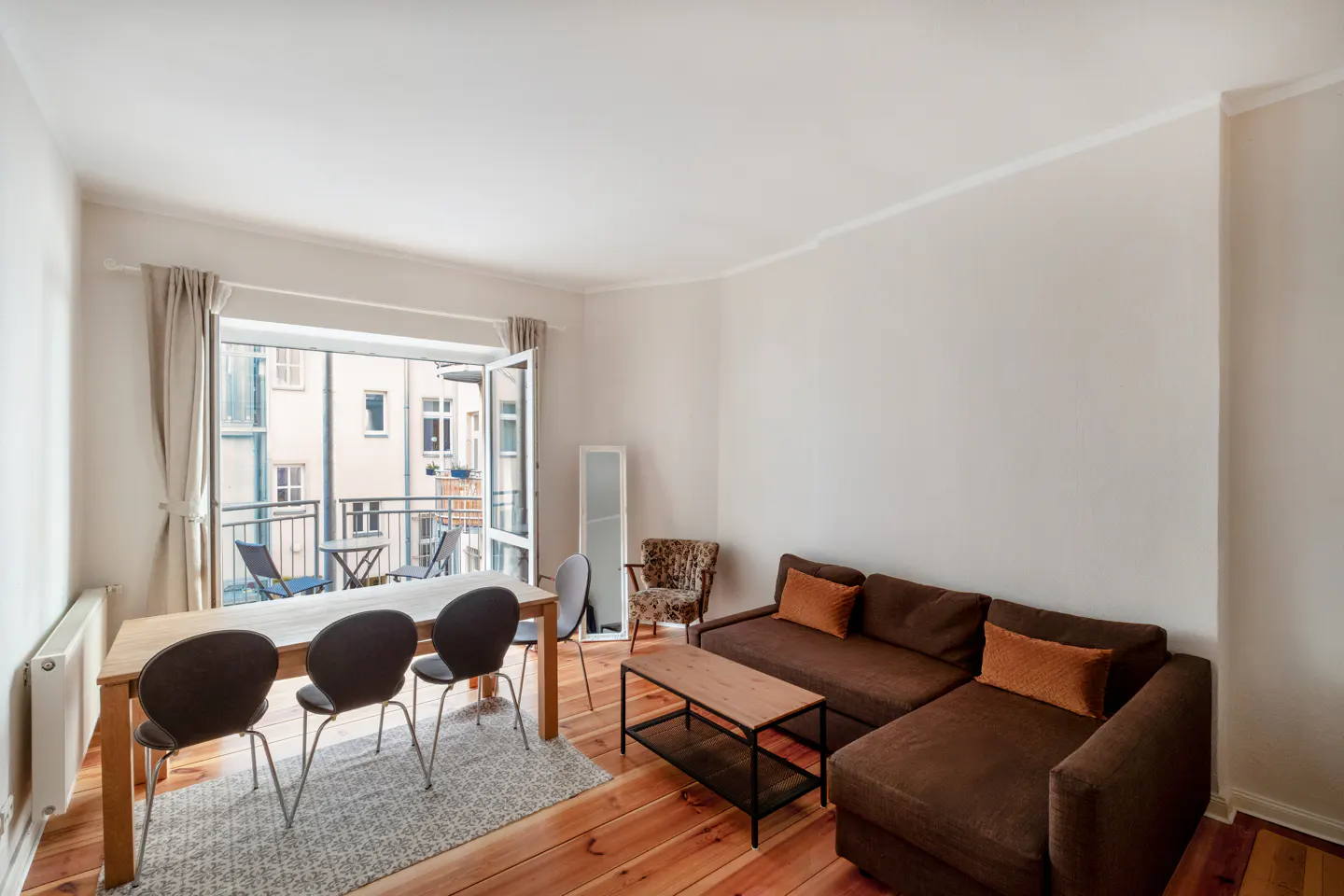 Bright living room with wood floors, brown sectional sofa, dining table, and balcony with outdoor seating.