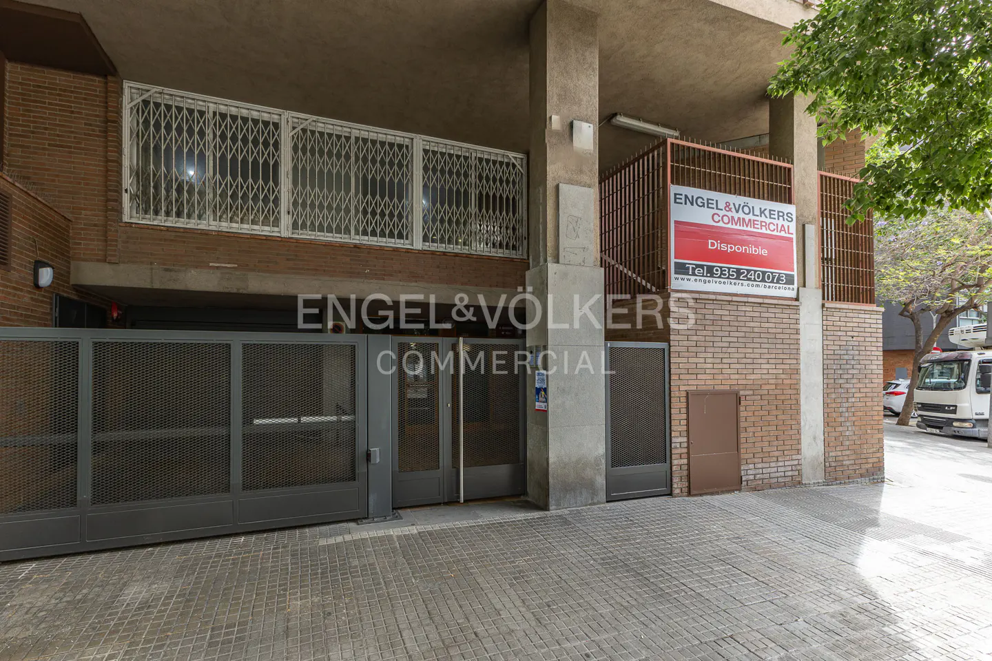 Exterior of a commercial building with an Engel & Völkers sign. The building has a gray gate and a brick facade. A truck is parked on the street.