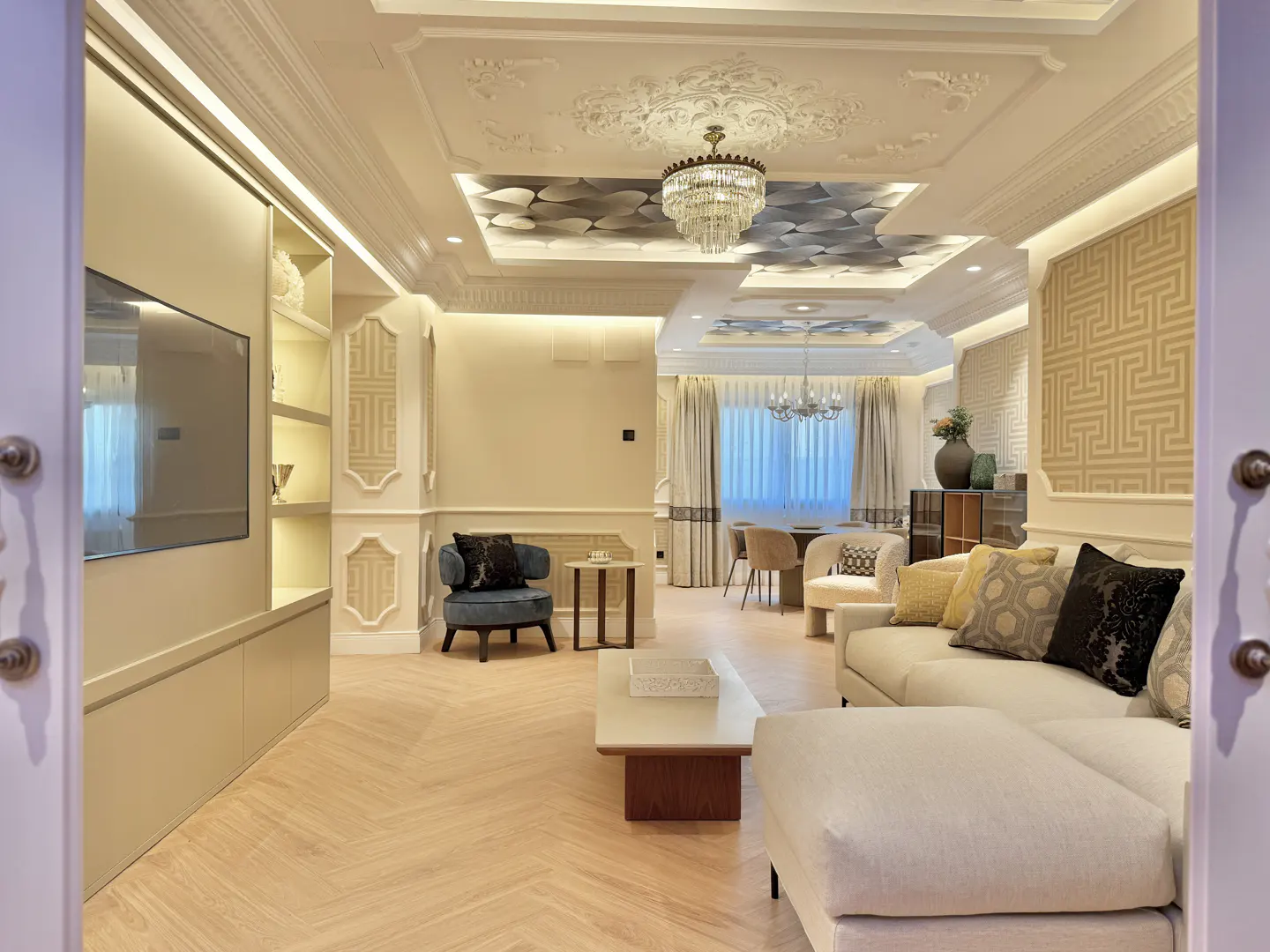 A bright, modern living room with a white sofa, blue chair, and herringbone wood floors. A chandelier hangs from the ornate ceiling.