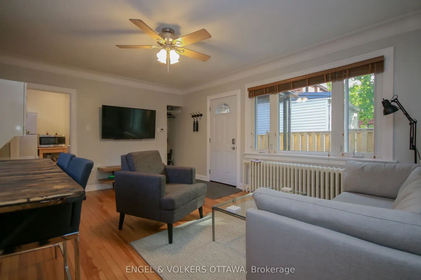 Living room with hardwood floors, gray sofa and chair, TV, ceiling fan, and large window with blinds.