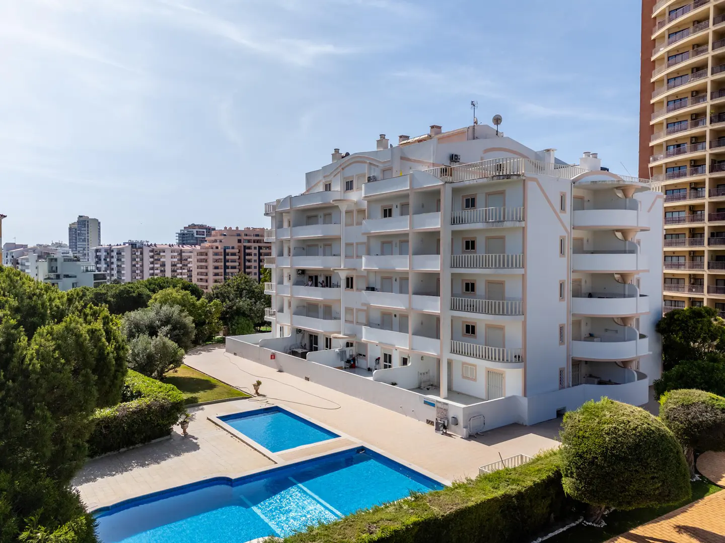 View of a white apartment building with balconies, two blue pools, green trees, and other buildings in the background.