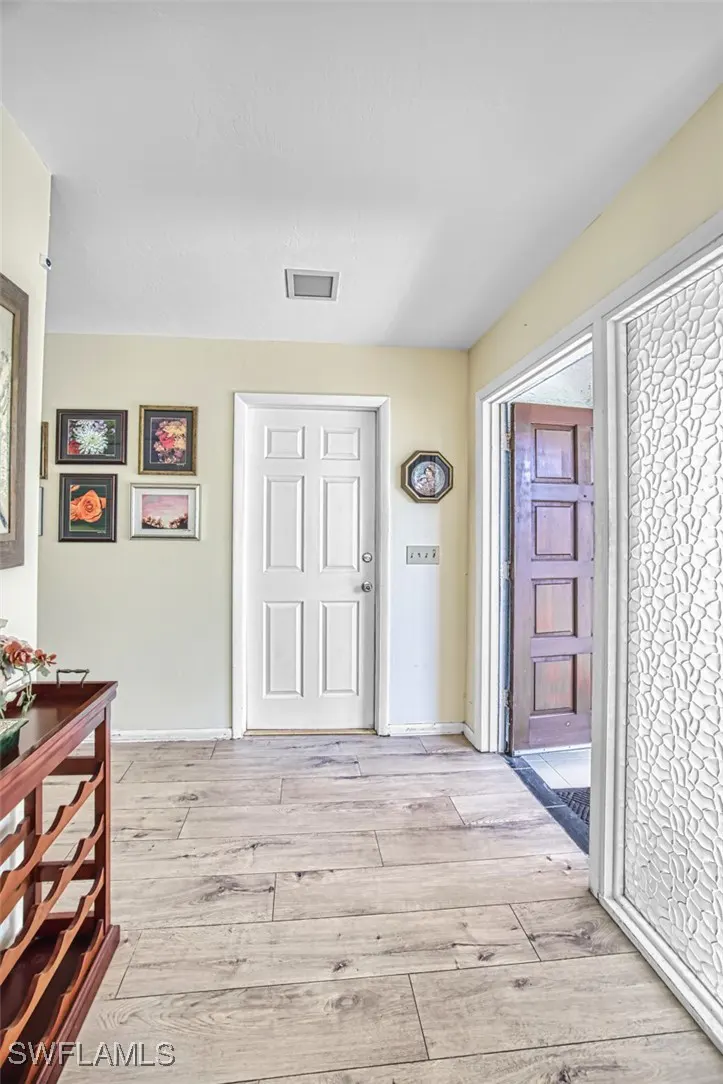 Entryway with light wood floors, white front door, and open brown door. A wine rack sits to the left, and framed art hangs on the wall.