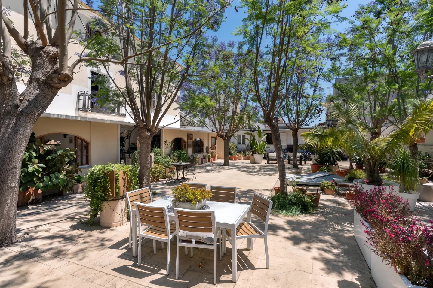 A sunny outdoor patio with a white dining set, surrounded by lush trees, potted plants, and a beige building.