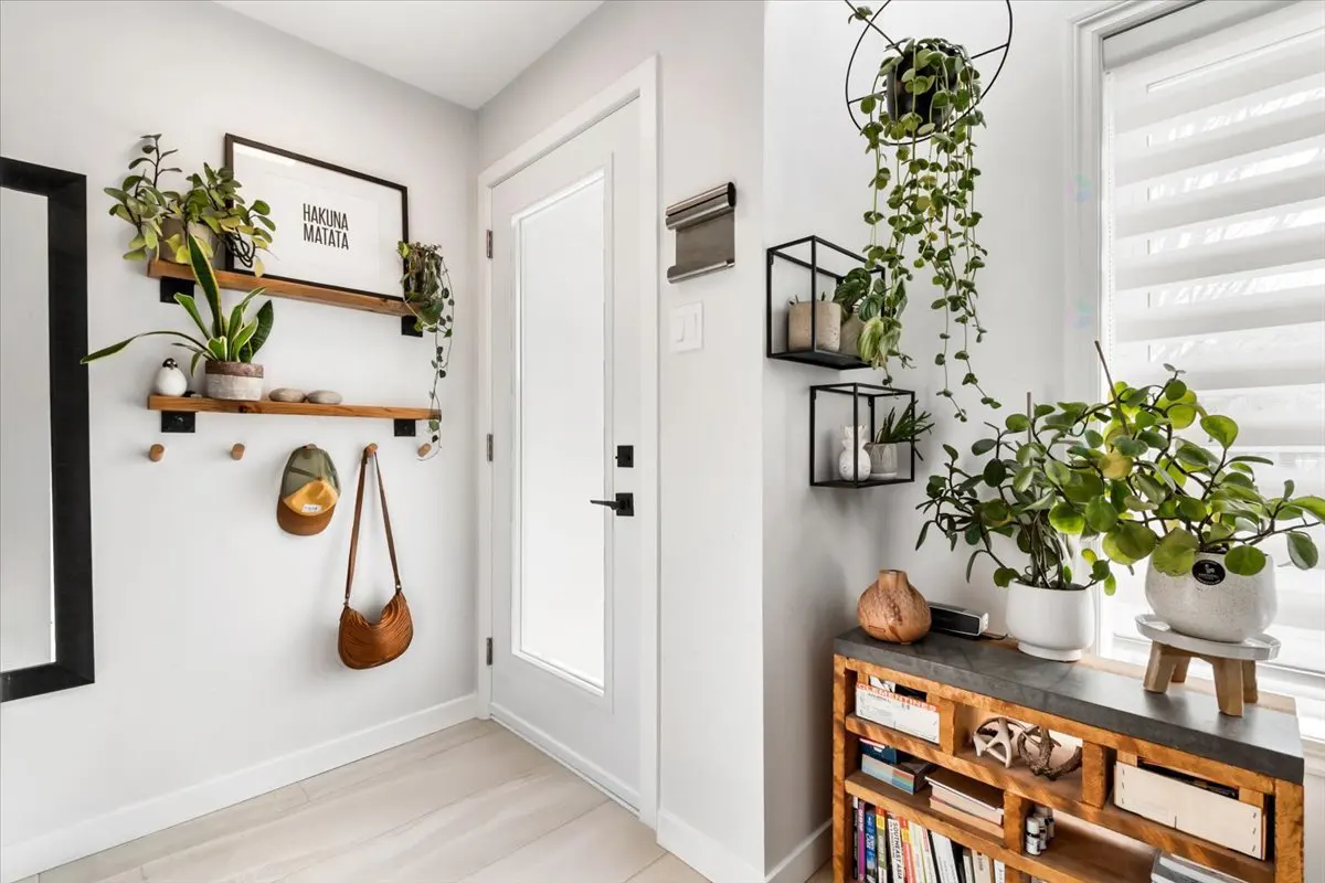 Bright entryway with white walls, light wood floors, and a white door. Shelves hold plants, a hat, and a bag. A wooden console table displays more plants.