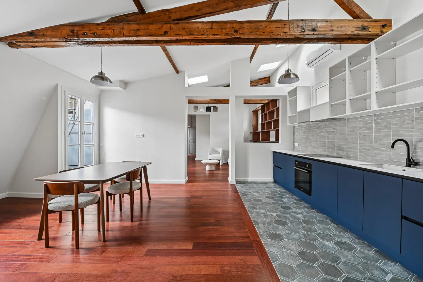Modern kitchen and dining area with exposed wooden beams, blue cabinets, grey hexagonal tile floor, and hardwood flooring.