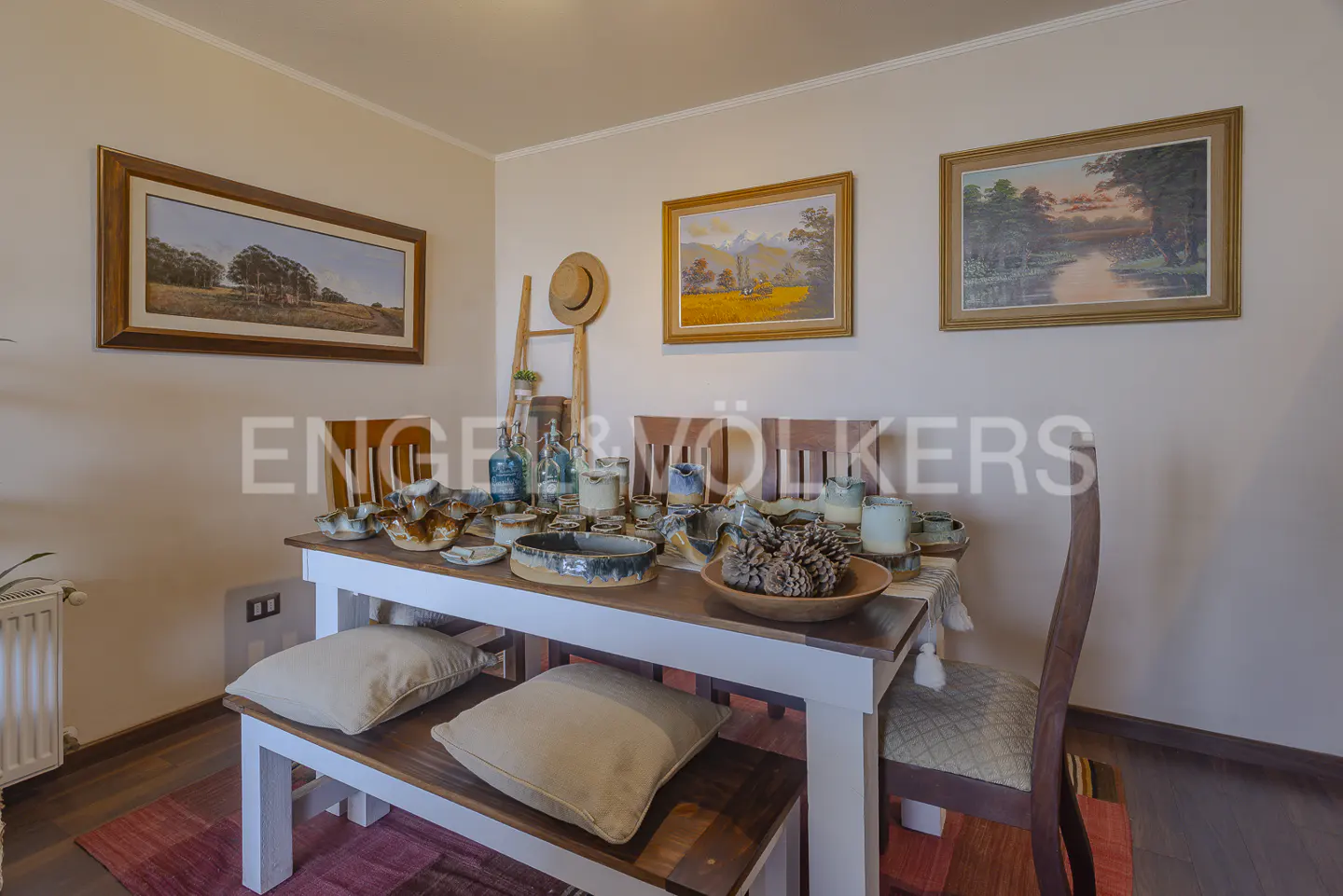 Dining room with a white table, wooden chairs, and a bench with pillows. Table is set with pottery and pinecones. Three landscape paintings hang on the wall.
