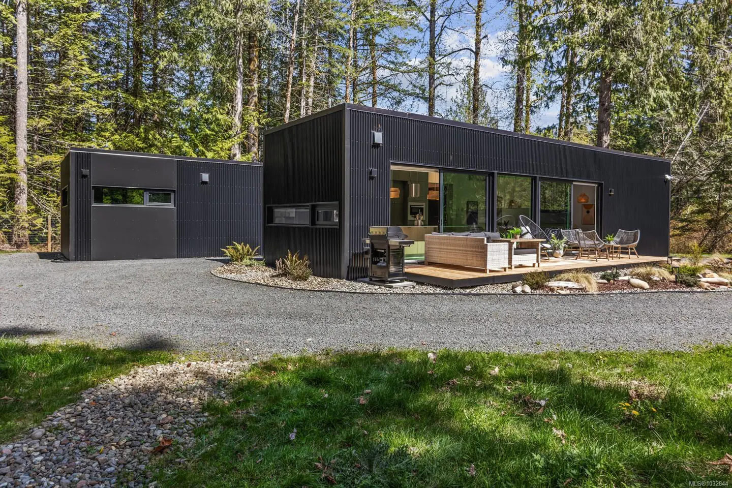 Modern black home with a deck and outdoor furniture, surrounded by gravel, grass, and trees.