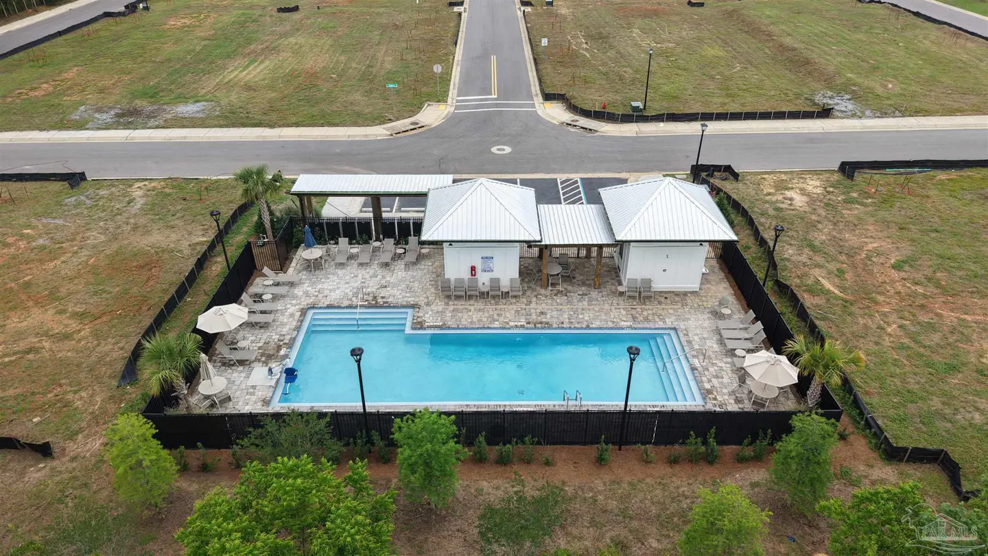 Aerial view of a community pool with blue water, lounge chairs, and white cabanas, surrounded by a black fence and green landscaping.