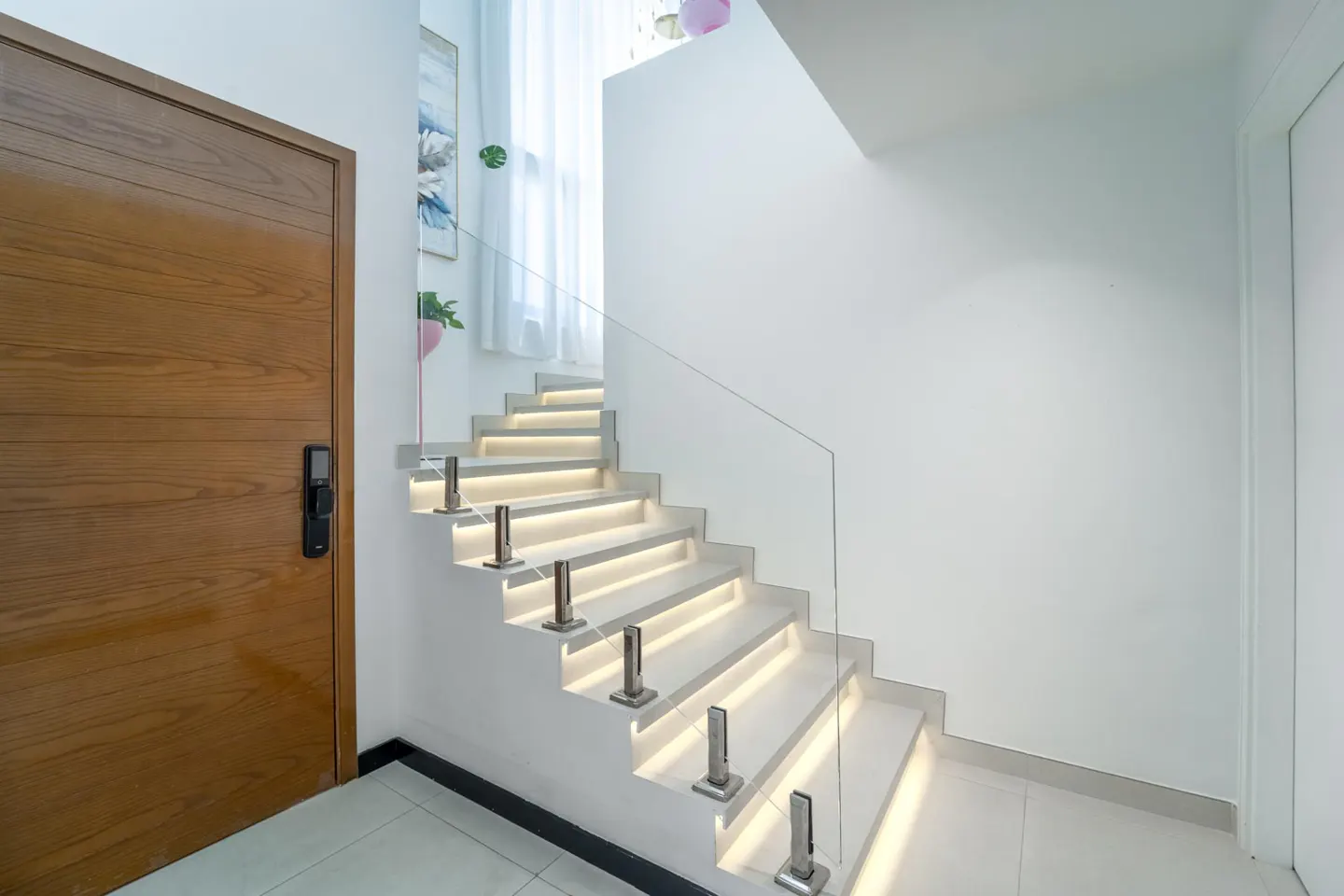 Modern home interior with white stairs, glass railing, and LED lighting. A wooden door is on the left, and white walls surround the staircase.