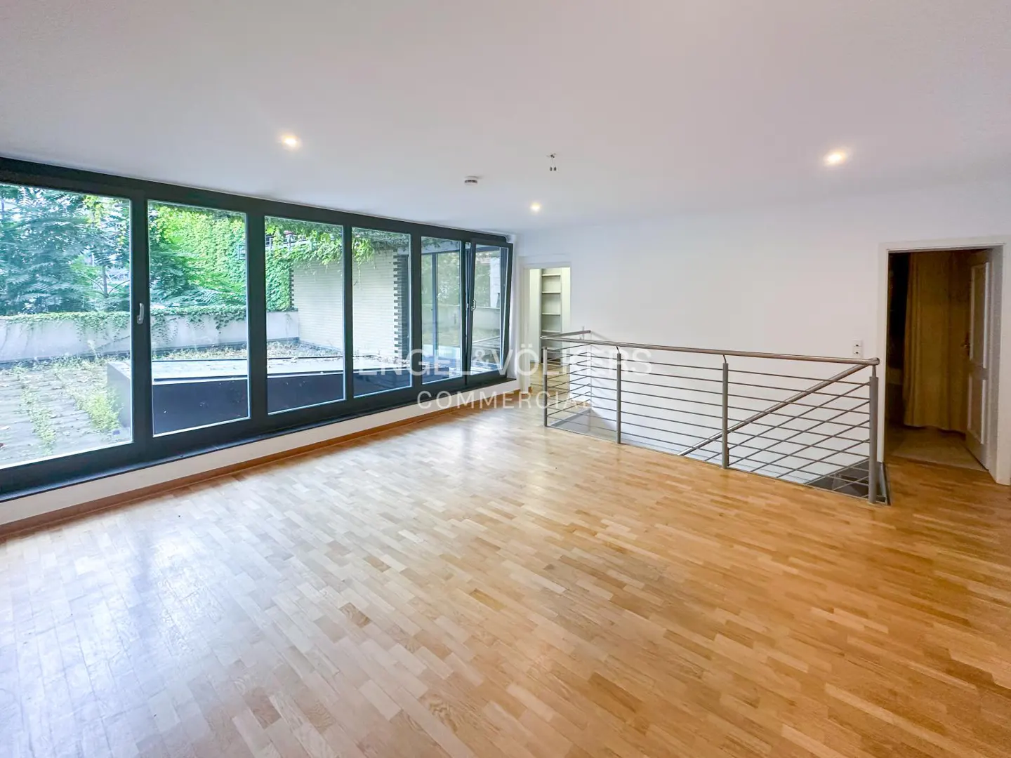 Bright, empty room with wood floors, large windows overlooking greenery, and a metal railing overlooking a lower level.