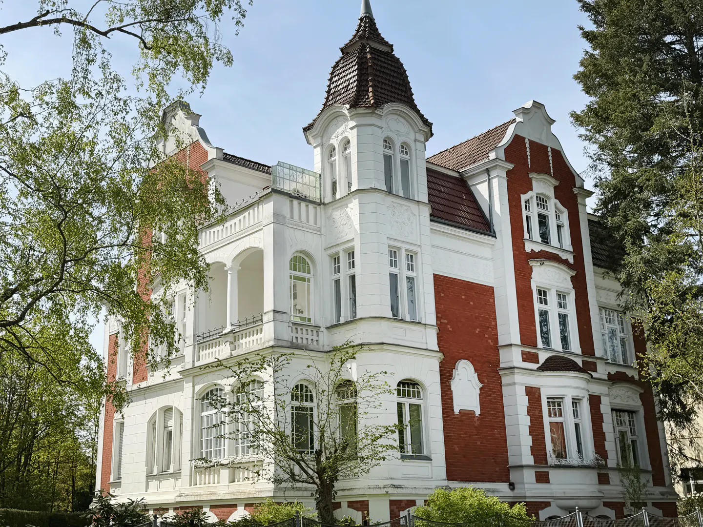 A three-story house with white walls and red brick accents, surrounded by trees.