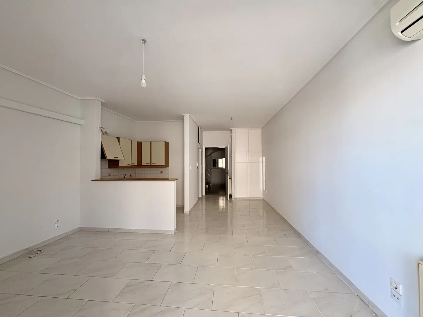 Bright, open-plan living area with white walls, tiled floors, a small kitchen with wood cabinets, and a white closet.