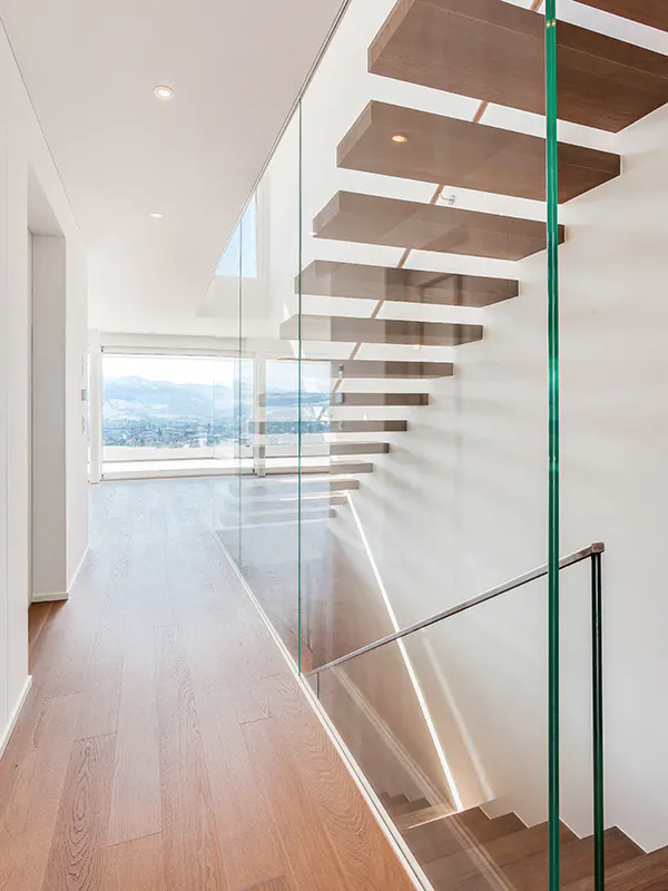Modern home interior with floating wood stairs, glass railings, and hardwood floors. Large windows offer a scenic view.
