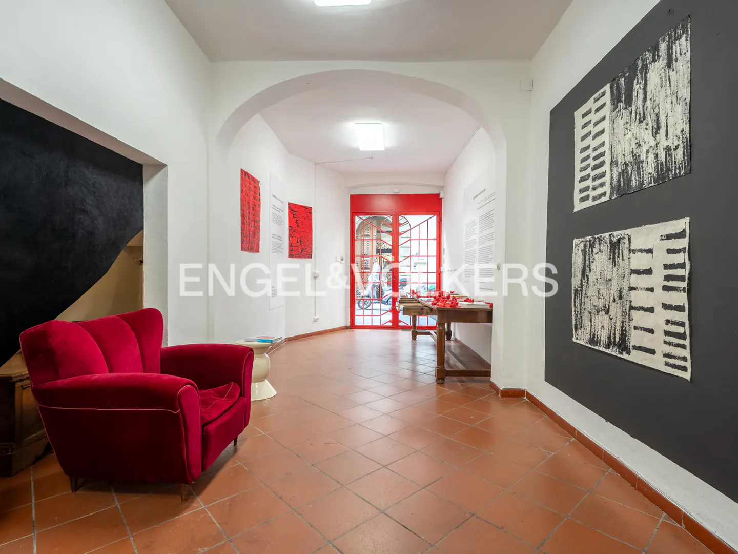A bright gallery space with a red armchair, terracotta tile floors, abstract art on walls, and a red-framed glass door.