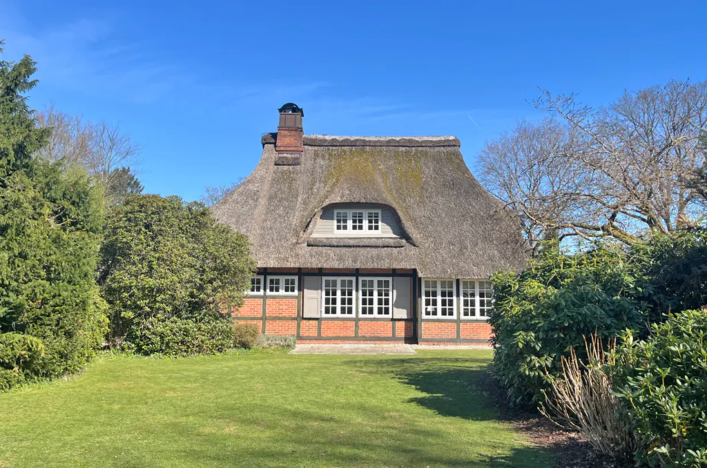 Charming thatched-roof cottage with red brick accents, surrounded by lush green lawn and trees under a clear blue sky.