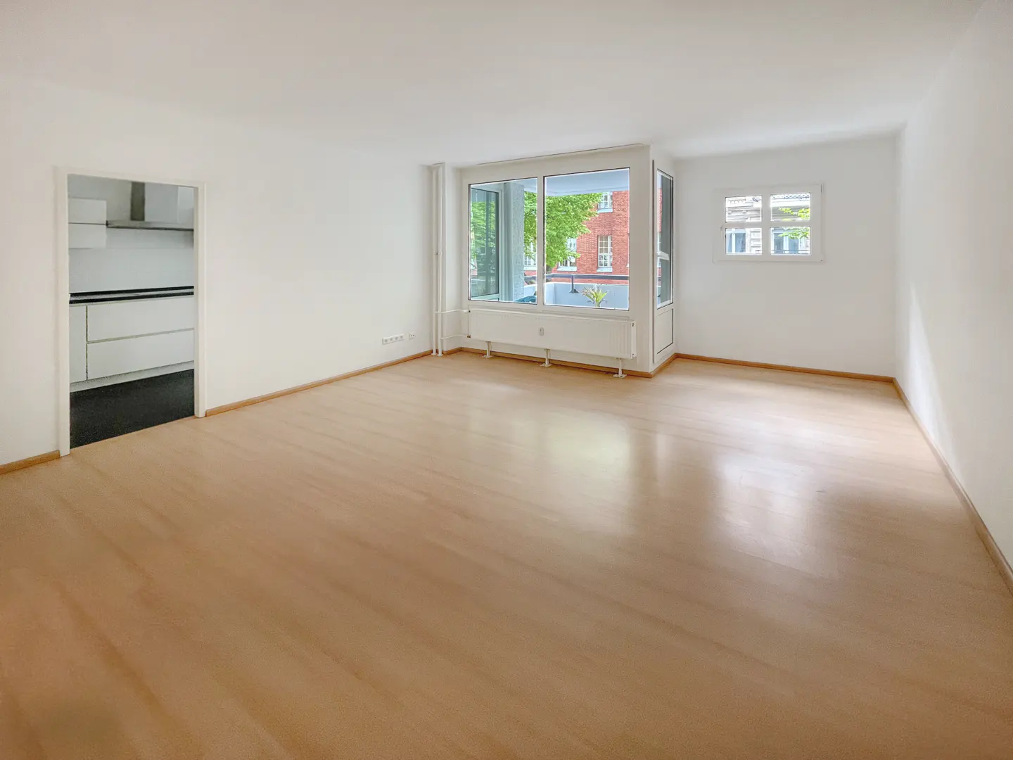 Bright, empty room with light wood floors and white walls. A doorway leads to a kitchen. Large windows offer a view of greenery and a brick building.