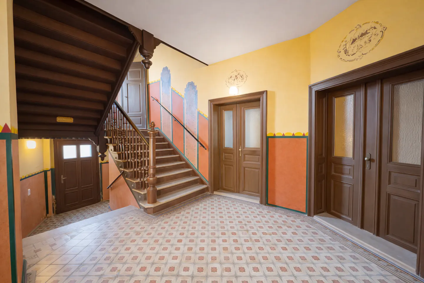 A bright hallway with patterned tile floors, a wooden staircase, and yellow walls featuring decorative painted accents.