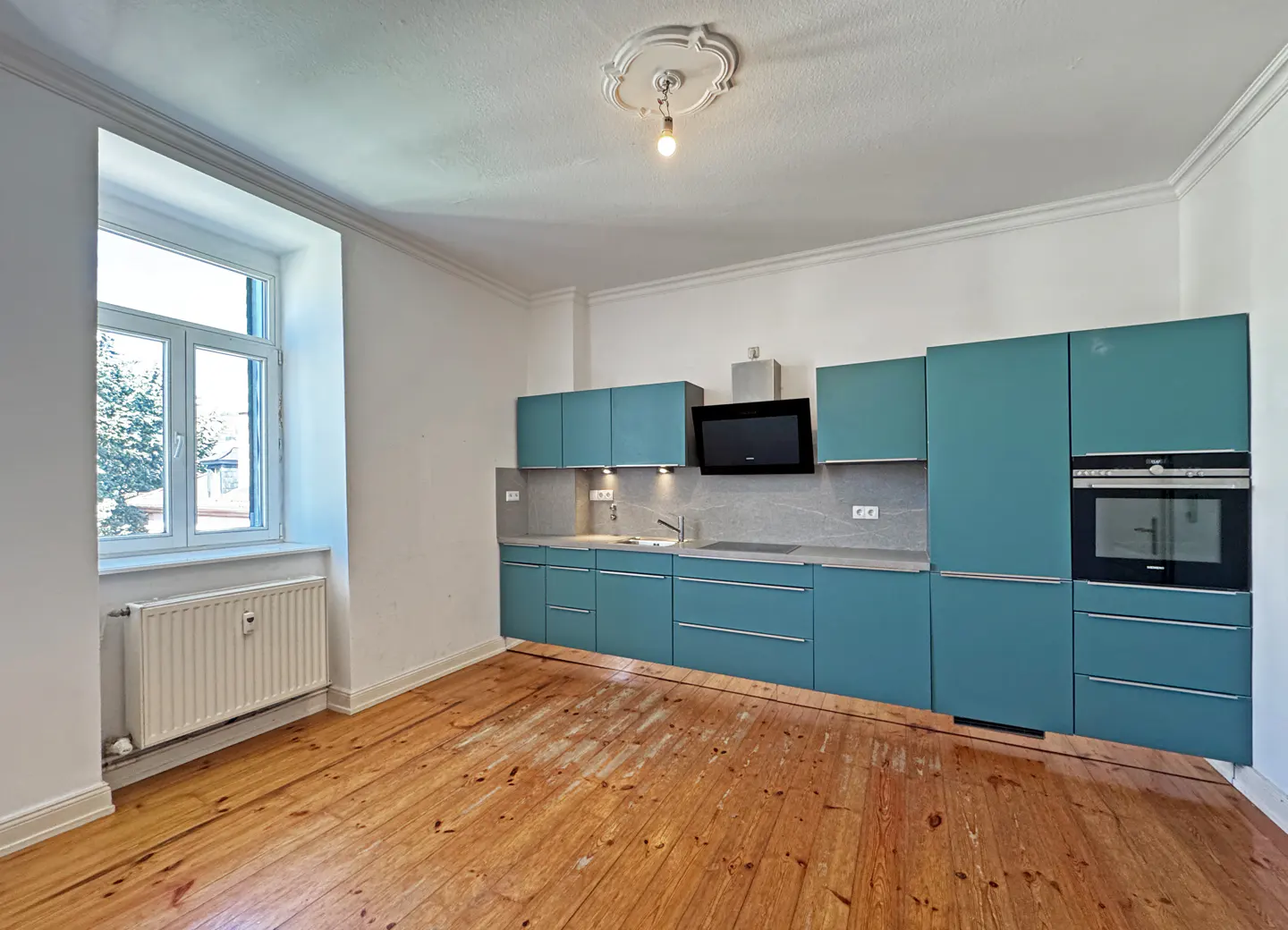 Bright kitchen with teal cabinets, gray countertops, and wood floors. A window provides natural light.