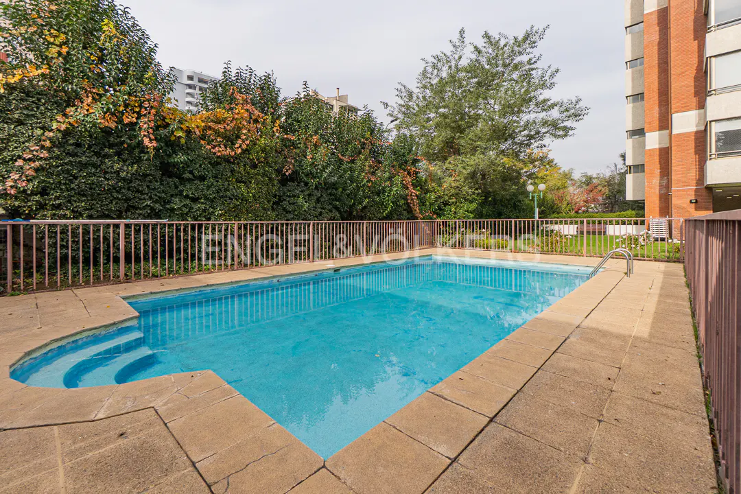 Outdoor swimming pool with blue water, stone deck, and metal fence, bordered by lush green trees and a brick apartment building.