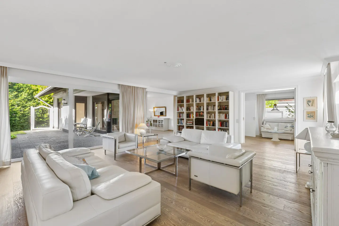 Bright living room with white sofas, a glass coffee table, and a large bookcase filled with books. Sliding glass doors lead to an outdoor patio.