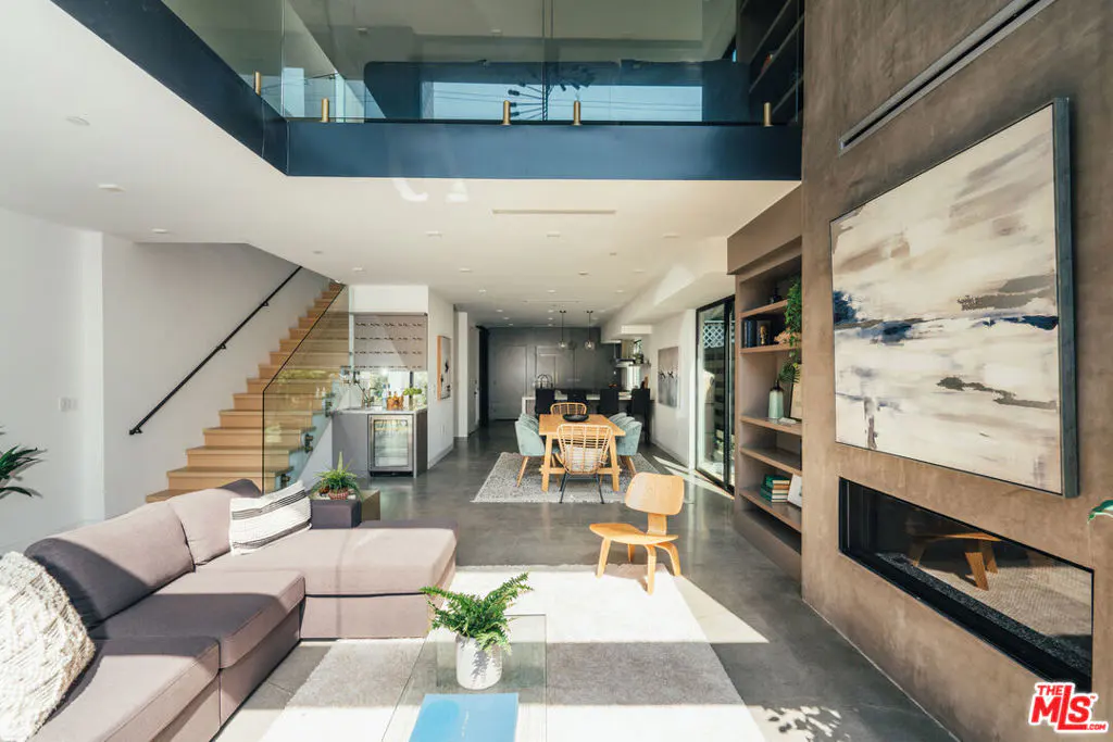 Open-concept living space with a gray sofa, wood stairs, and a dining area. A large painting hangs above a modern fireplace.