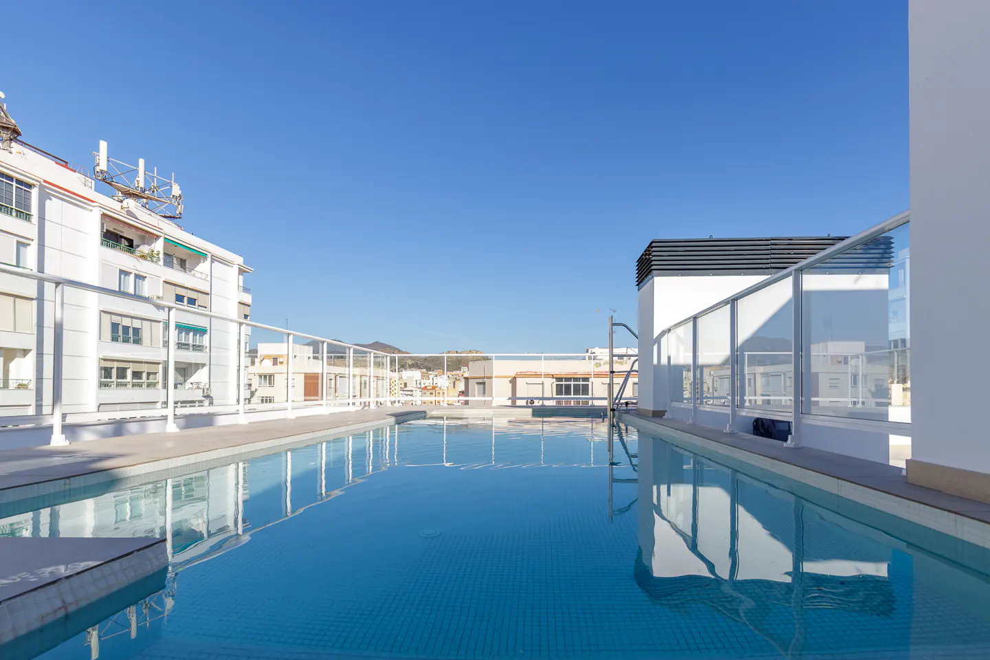Rooftop pool with clear blue water, surrounded by white railings and buildings under a clear blue sky.