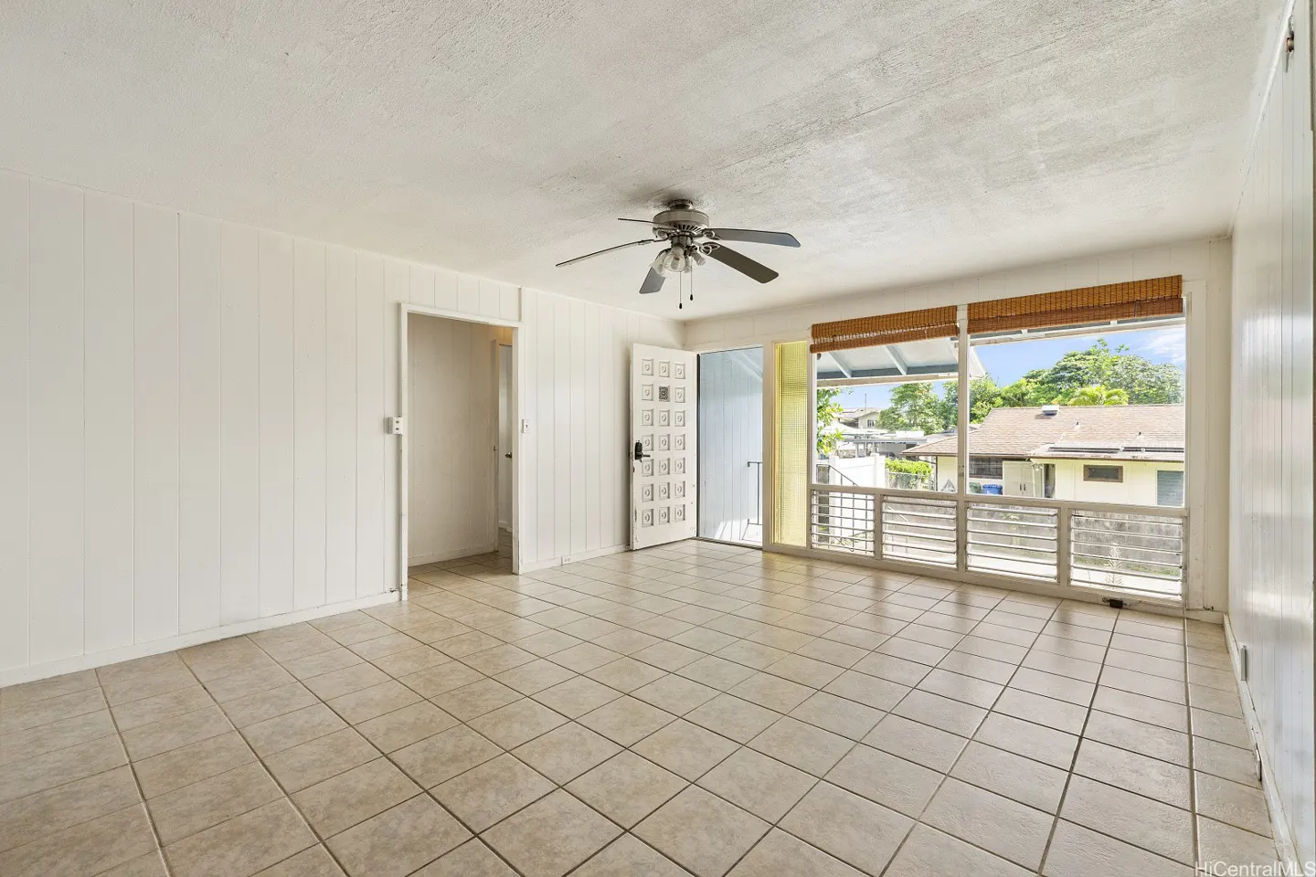 Empty living room with white walls, tiled floors, a ceiling fan, and a large window looking out to a residential street.