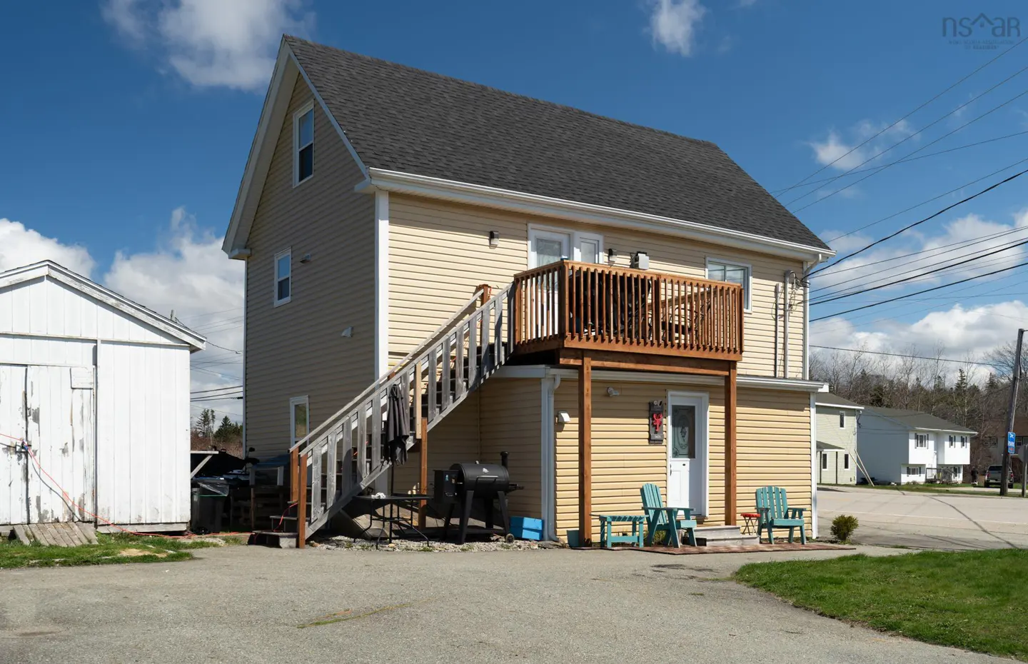 Two-story tan house with a dark roof, wooden deck, and stairs. A white shed is on the left, and blue chairs sit outside the lower door.