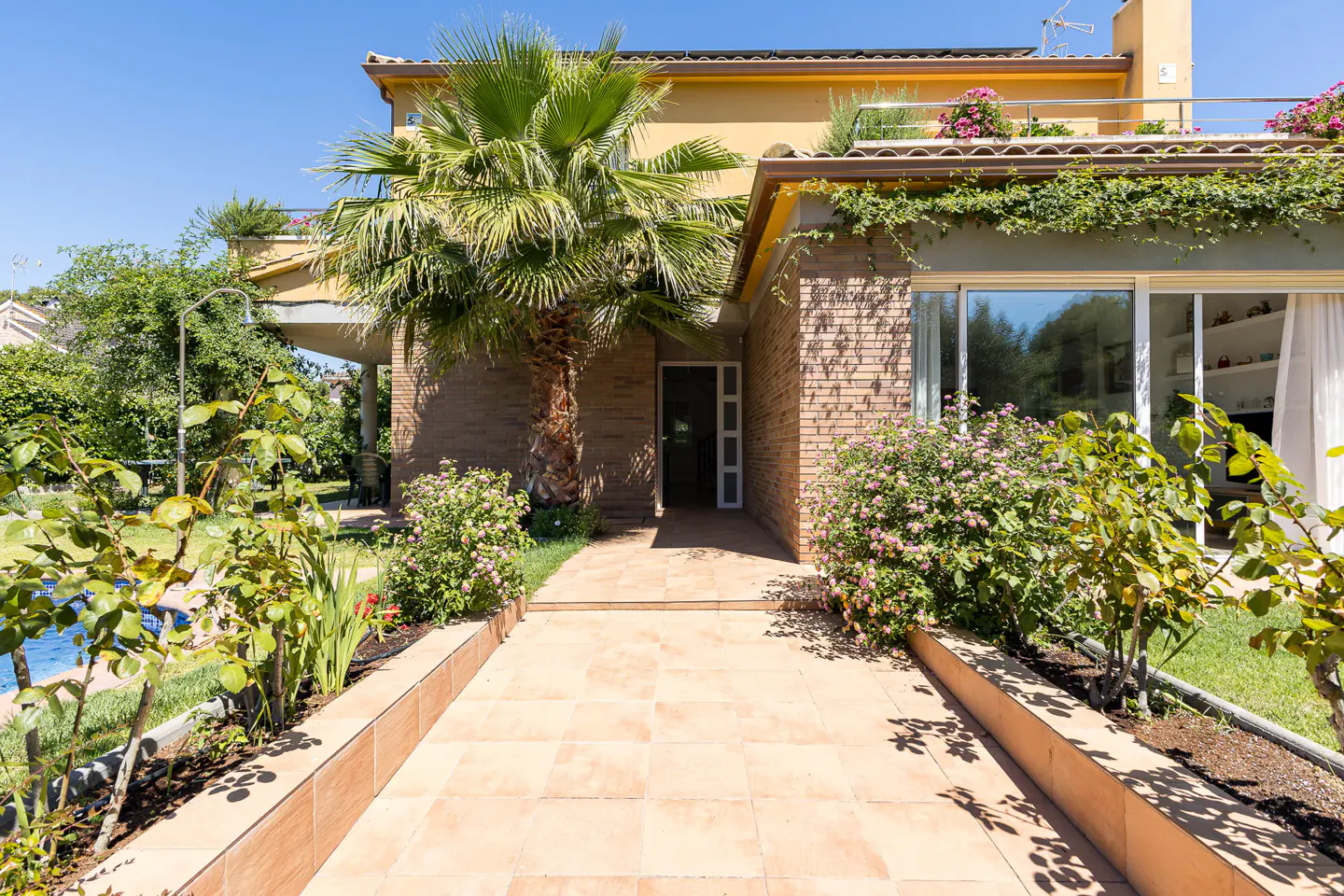 Tiled walkway leading to a brick house entrance with a large palm tree, lush green plants, and pink flowers.