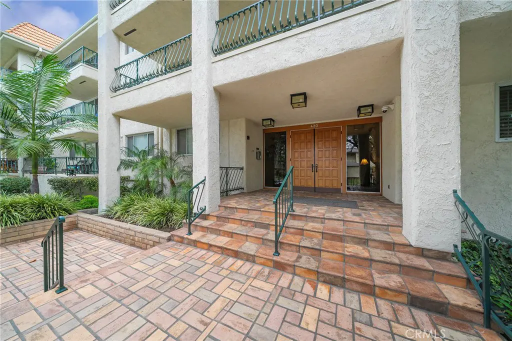 Exterior view of a white apartment building with brown double doors and brick steps leading to the entrance. Green plants and palm trees surround the building.